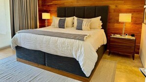Un dormitorio con una cama grande, cabecera de madera, dos mesitas de noche con lámparas y una cortina gris.