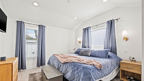Une chambre à coucher comprenant un lit, une table de chevet, un petit banc en bois, une commode, un téléviseur fixé au mur et une fenêtre avec des rideaux.