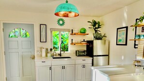 A compact kitchen with white cabinets, a stainless steel refrigerator, a microwave, and a sink. There is a window with a view of greenery outside, and a teal pendant light hangs above the counter. - Ho’okipa Hideaway - North Shore, Walk to Cafe (Haiku-Pauwela)