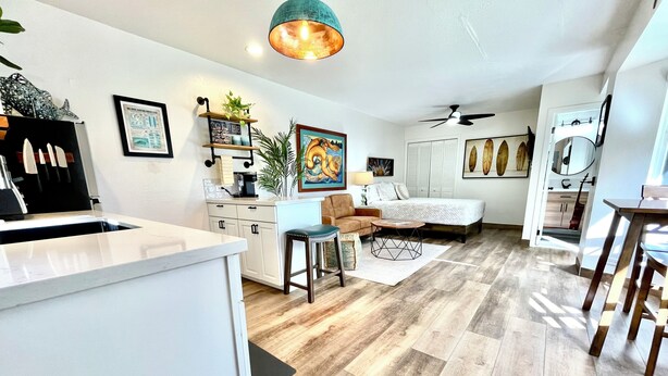 A modern kitchen with white cabinets and a central island, a cozy seating area with a sofa and coffee table, and a bedroom with a bed and a ceiling fan. - Ho’okipa Hideaway - North Shore, Walk to Cafe (Haiku-Pauwela)