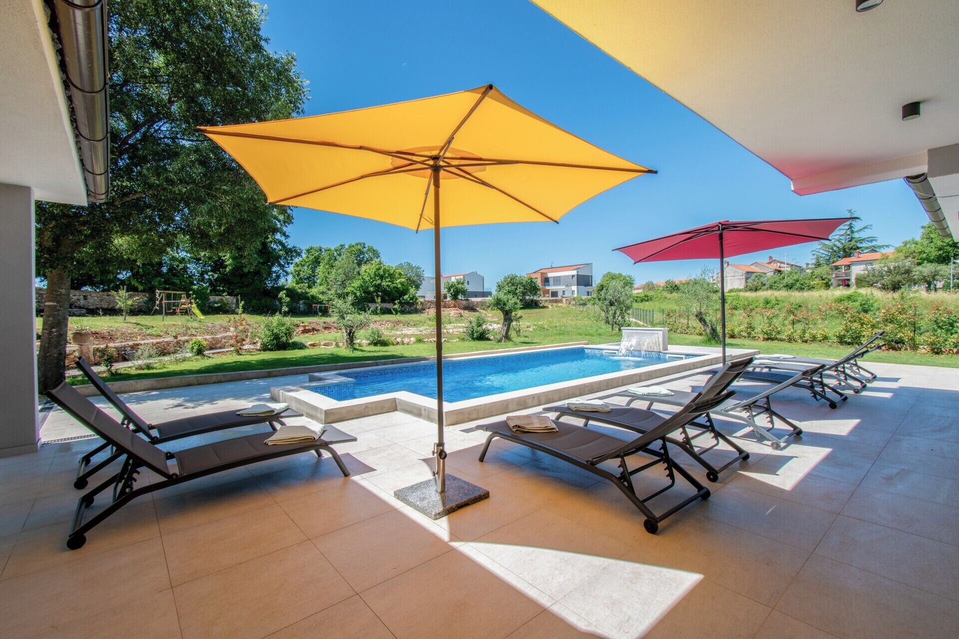 Un espace aménagé au bord de la piscine, avec deux chaises longues et deux parasols, offrant une vue dégagée sur la piscine et donnant sur un jardin paysager en arrière-plan.