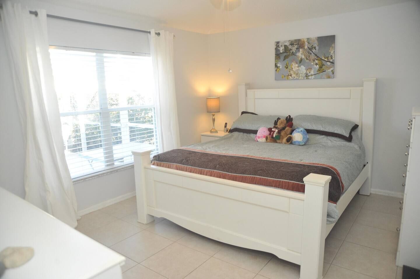 A bedroom with a bed, a nightstand, a lamp, a window with curtains, and a painting on the wall.