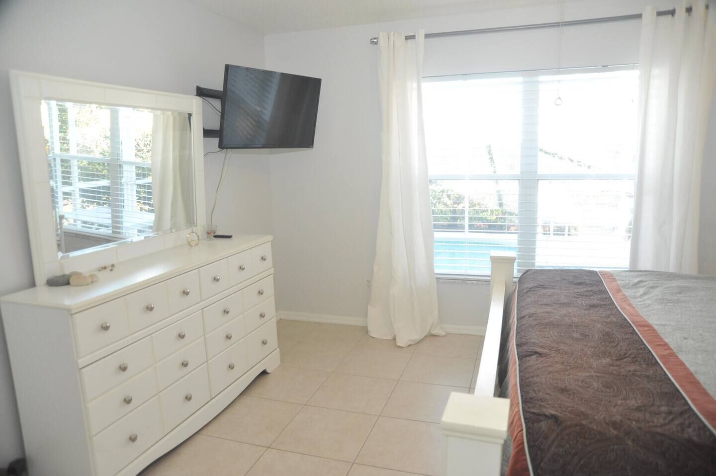 A bedroom with a bed, a dresser, a television mounted on the wall, and a window with curtains.