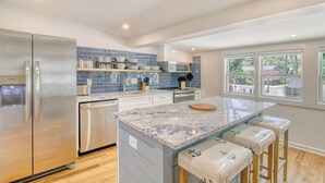 Una cocina moderna con un refrigerador de acero inoxidable, un lavavajillas, una isla de mármol con taburetes y vistas al exterior a través de amplias ventanas.