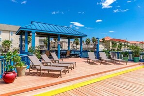 Terraza de madera con sillones de descanso y un pabellón cubierto, rodeada de edificios y palmeras.