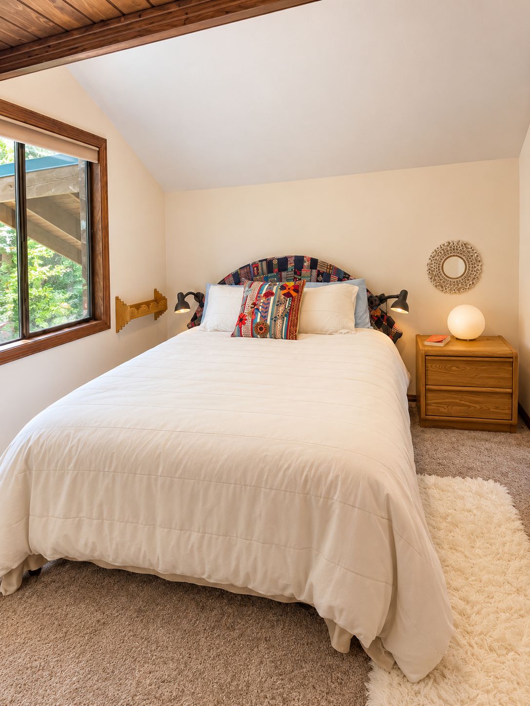 A bedroom with a large bed, a nightstand with a lamp, a decorative mirror, and a window with a view of greenery.