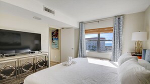 Une chambre d’hôtel avec un grand lit, un téléviseur à écran plat posé sur un meuble, une fenêtre avec des rideaux et un tableau accroché au mur.