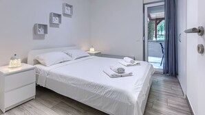 A modern bedroom with a large bed, white nightstands, a bedside lamp, and a view of the outdoors through a sliding glass door.