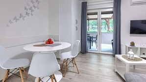 A modern living room with a round white table, white chairs, a flat-screen TV, and a sliding glass door leading to an outdoor area.