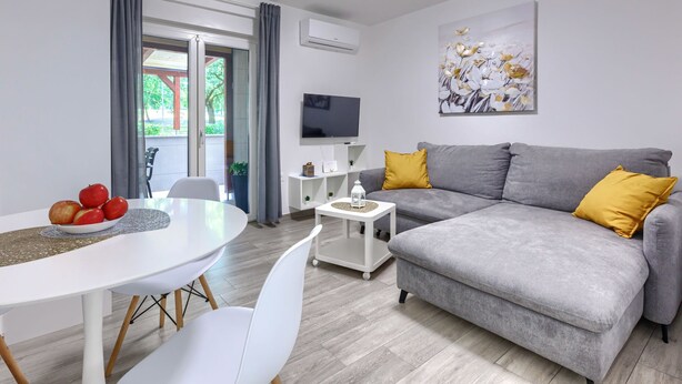 A modern living room with a grey sofa, white chairs, a dining table with fruit, a TV, and a painting.