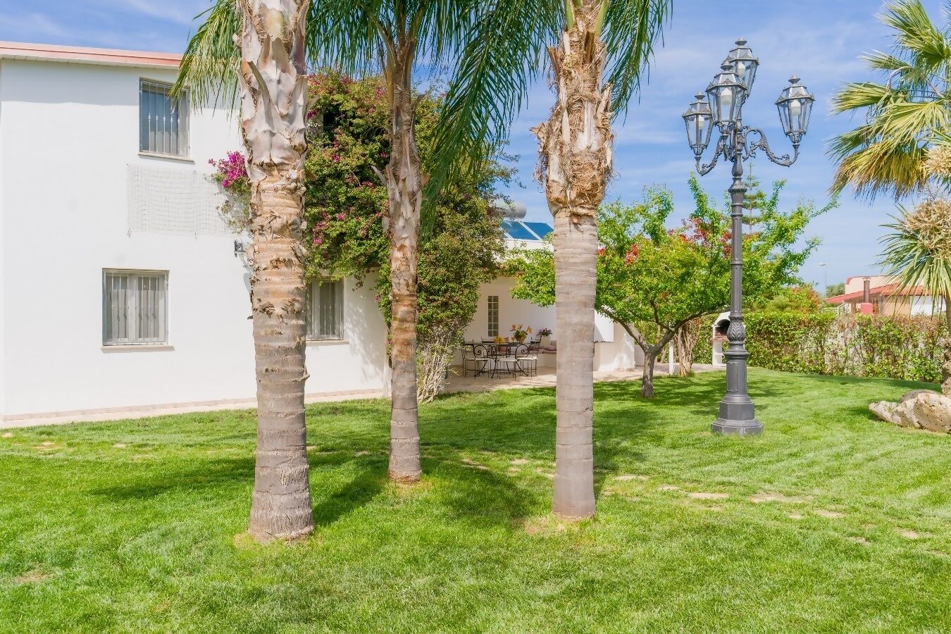 Un jardin bien entretenu, agrémenté de palmiers, avec un espace repas et un bâtiment blanc doté de fenêtres.