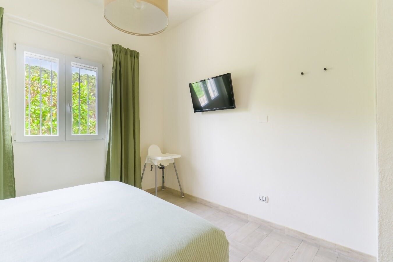 Un dormitorio con cama, televisión montada en la pared, ventana con cortinas y una mesita.