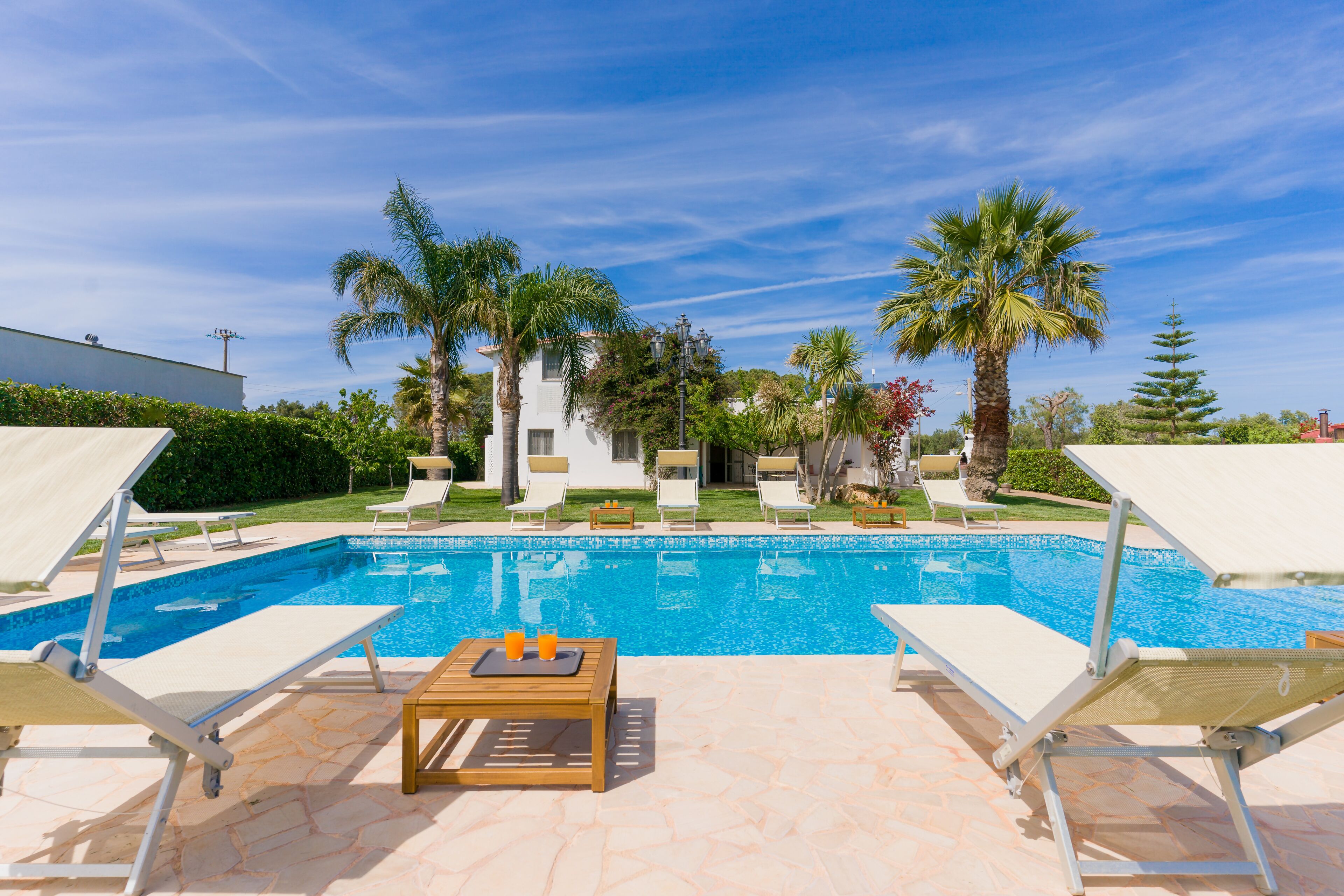 Ein Swimmingpool mit Liegestühlen und einem Tisch, umgeben von Palmen und blauem Himmel.