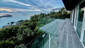 Un balcon doté d’une balustrade en verre, donnant sur une zone côtière avec des bateaux et de la végétation.