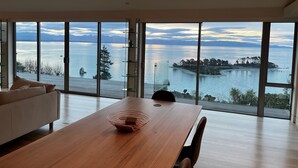 Un intérieur moderne avec une grande table en bois et un canapé blanc, de grandes fenêtres offrant une vue sur un lac et des montagnes lointaines.