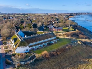 Un grand bâtiment blanc, doté d’un toit de chaume, entouré d’une zone herbeuse et bordé par une étendue d’eau.
