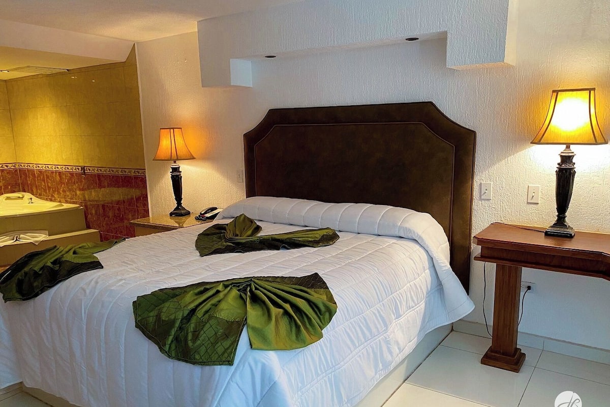 Une chambre d’hôtel avec un grand lit, deux lampes de chevet, une table de chevet en bois, une petite table et une étagère fixée au mur.