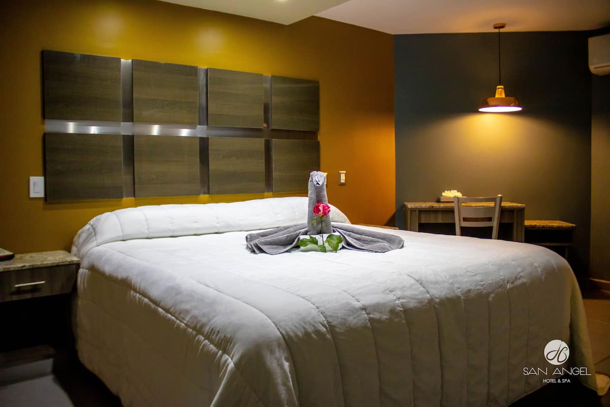 Une chambre d’hôtel avec un grand lit, une tête de lit ornée d’un panneau décoratif, une petite table avec une chaise et un luminaire suspendu.