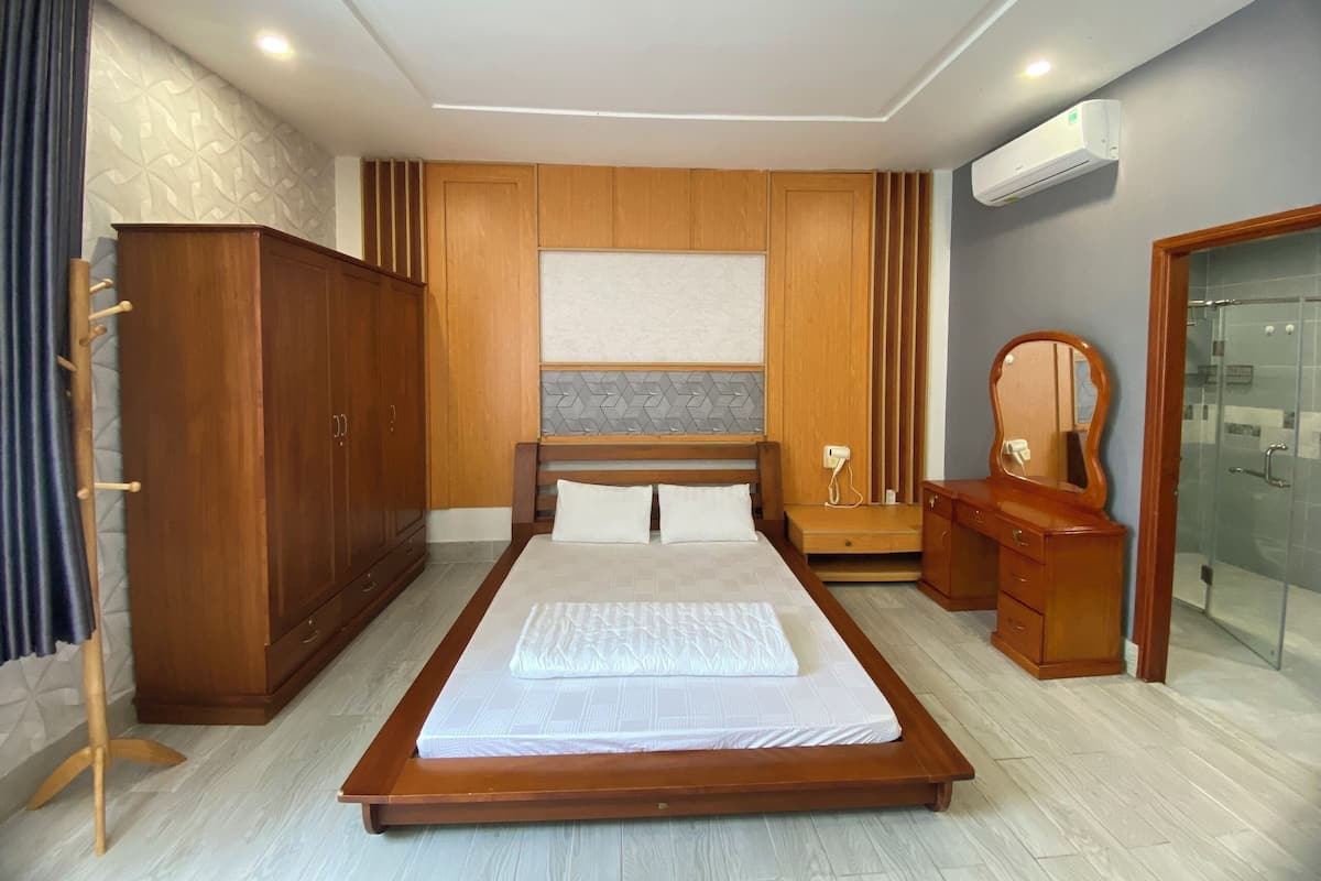 A bedroom with a large bed, wooden wardrobe, vanity with mirror, and built-in air conditioning unit.