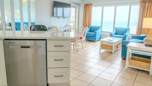 Ein modernes Wohnzimmer mit Flachbildfernseher, einer Essecke mit weißen Stühlen und einer Küche mit Geschirrspüler.