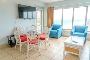 A room with a dining table, four chairs, a TV mounted on the wall, two blue armchairs, and a view of the beach.