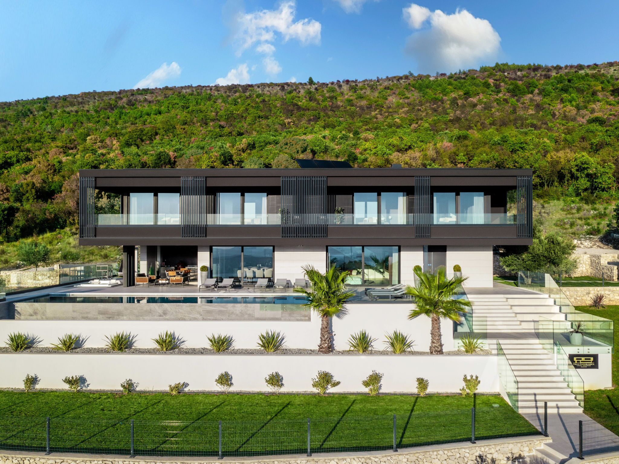 Une maison moderne à deux étages, dotée d’une piscine, entourée d’une pelouse bien entretenue et de palmiers.