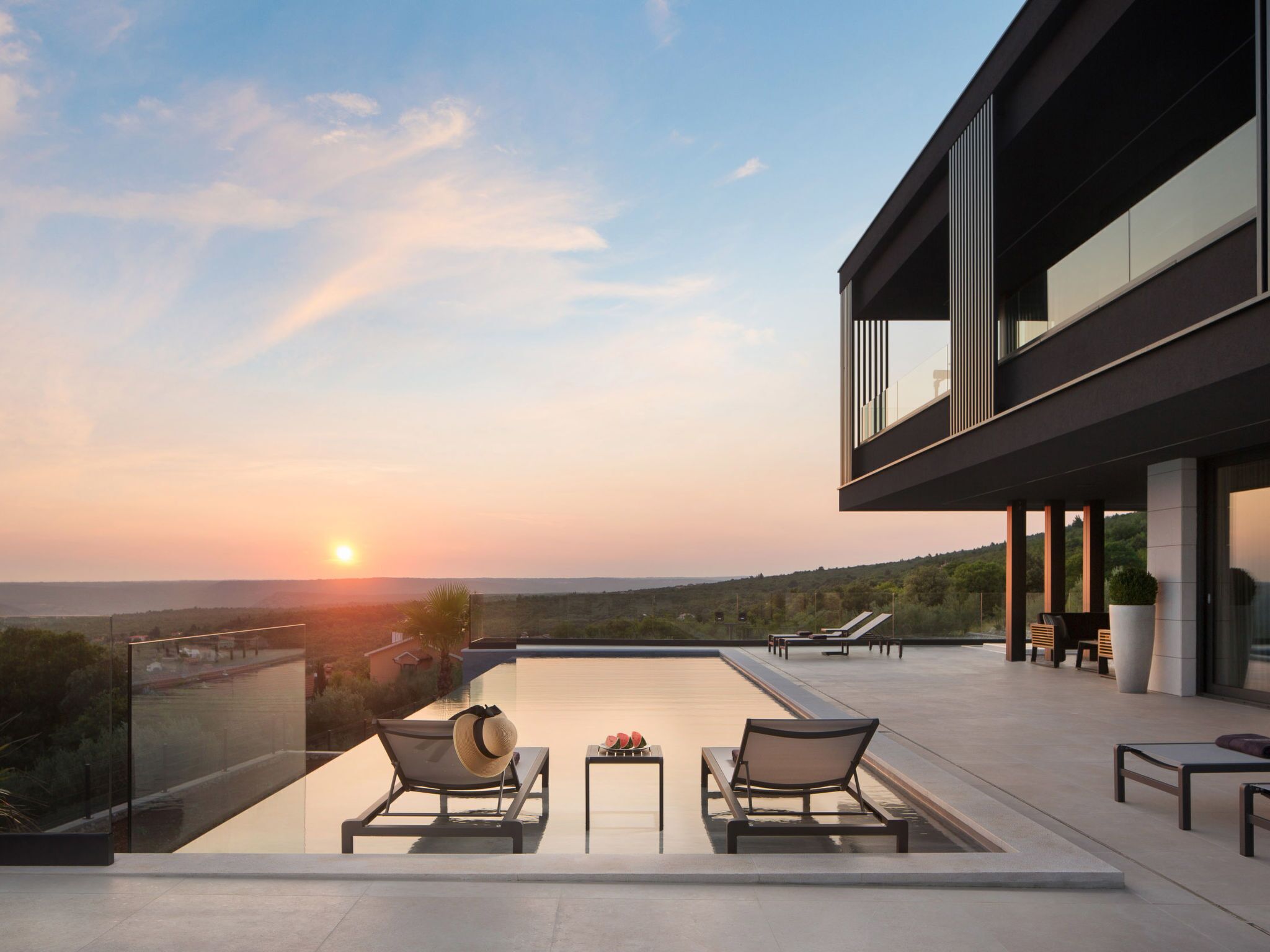 Une terrasse extérieure moderne dotée d’une piscine, de chaises longues et offrant une vue sur le coucher de soleil au-dessus d’un paysage vallonné.