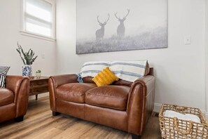A living room with a brown leather sofa, a blue and white patterned throw, and a yellow pillow. A large painting of deer is on the wall, and there is a wicker basket on the floor. - Square Haven Duplex -Walk to Pubs Cafe & Ice Cream (Washington)
