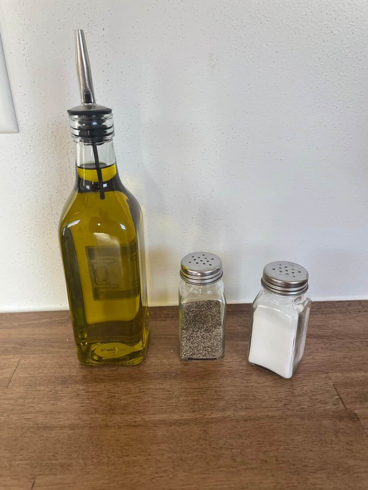 A bottle of olive oil, a salt shaker, and a pepper shaker on a wooden surface.