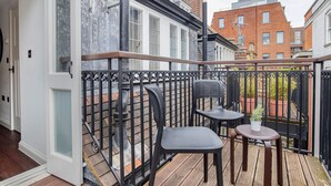En balkon med trægulv, gelænder i metal, to sorte stole og et lille bord med en potteplante.