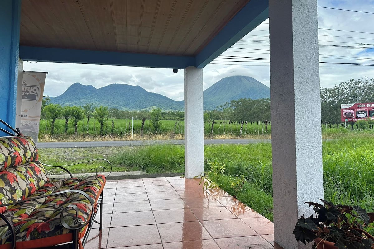 A covered patio with a bench, a view of a vineyard, and a mountain in the background.