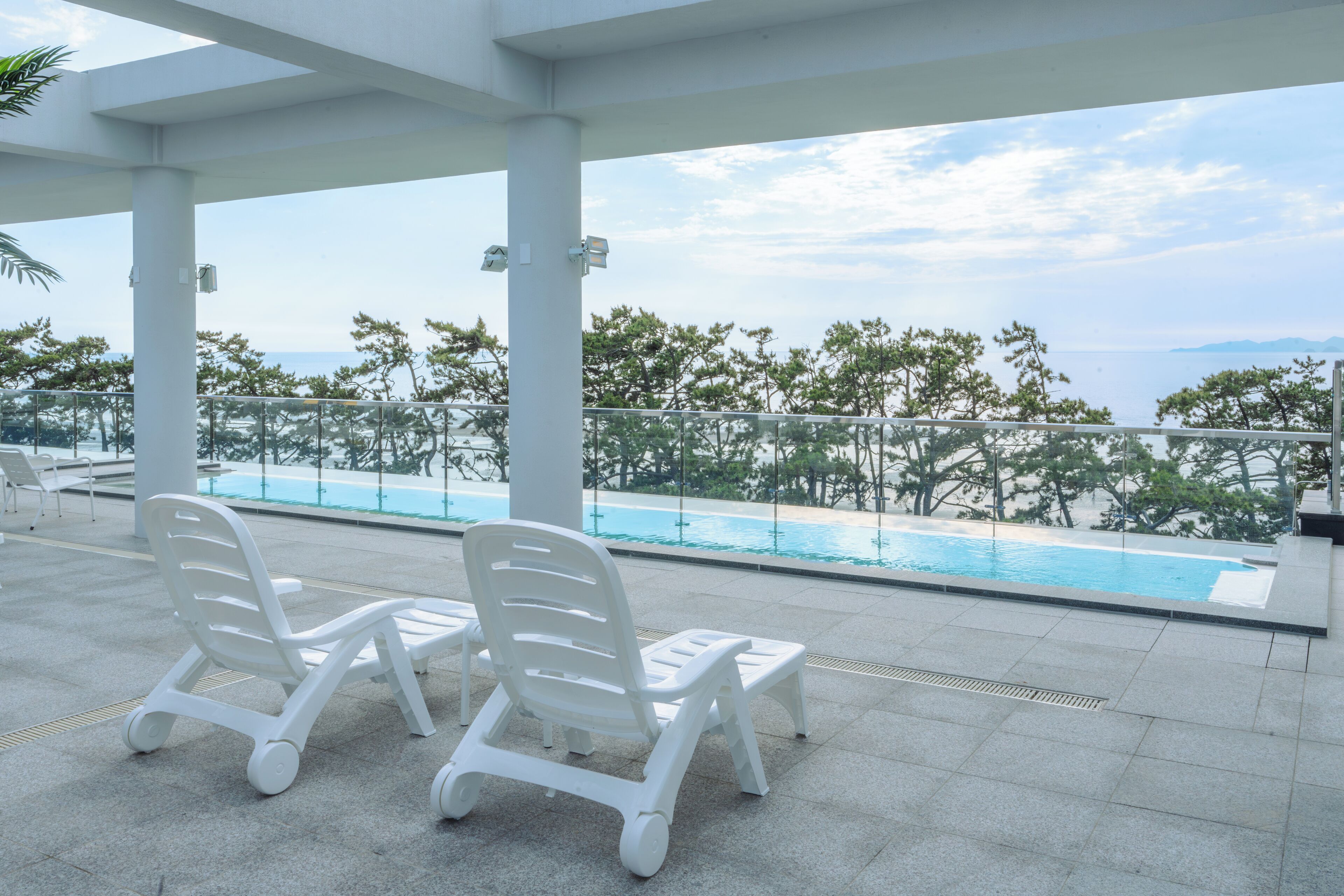 Un área de piscina en la azotea con dos sillones blancos y una vista despejada del mar y los árboles.