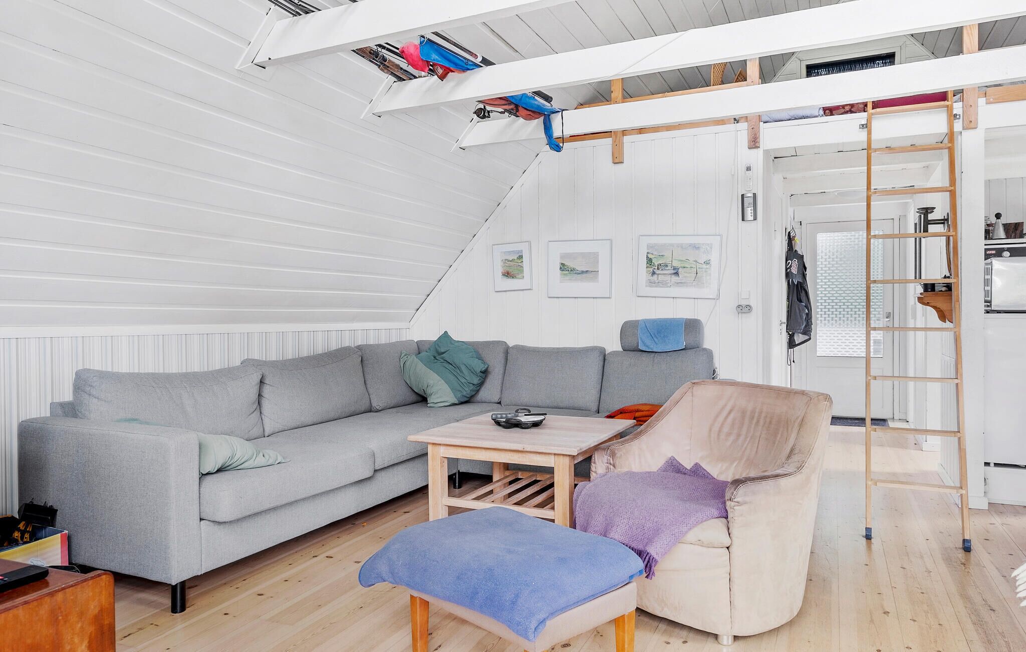 Ett rum i loftstil med en grå soffa, ett litet bord, en stol och en trästege. På väggen hänger inramade tavlor och det finns ett fönster med persienner.