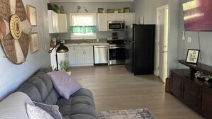 A modern kitchen with white cabinets, a black refrigerator, and stainless steel appliances. There is a grey sofa with a patterned throw pillow in the living area.