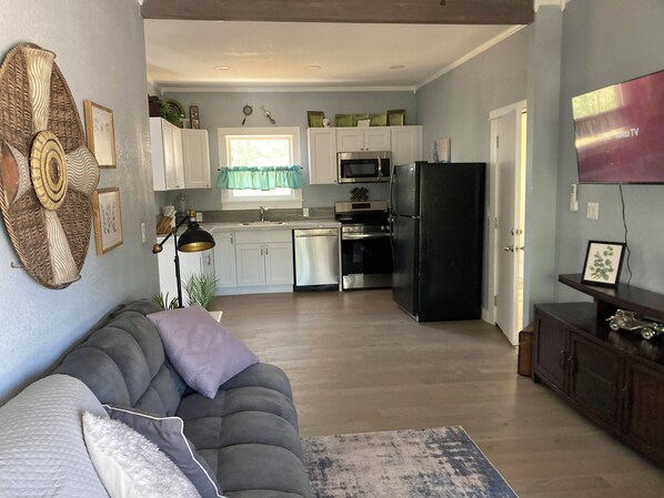 Een moderne keuken met witte kasten, een zwarte koelkast en roestvrijstalen apparatuur. In de woonkamer staat een grijze bank met een kussen met een patroon.