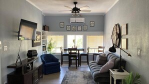 A living room with a sofa, a blue armchair, a dining table with chairs, a ceiling fan, and a TV mounted on the wall.