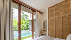 A bedroom with a bed, a wooden wardrobe, a vanity with a mirror, and a view of a swimming pool. - Gaia Villa by Betterplace (Canggu)