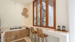 A modern kitchen with a central island, two bar stools, a window with a view, and decorative plates on the wall. - Gaia Villa by Betterplace (Canggu)