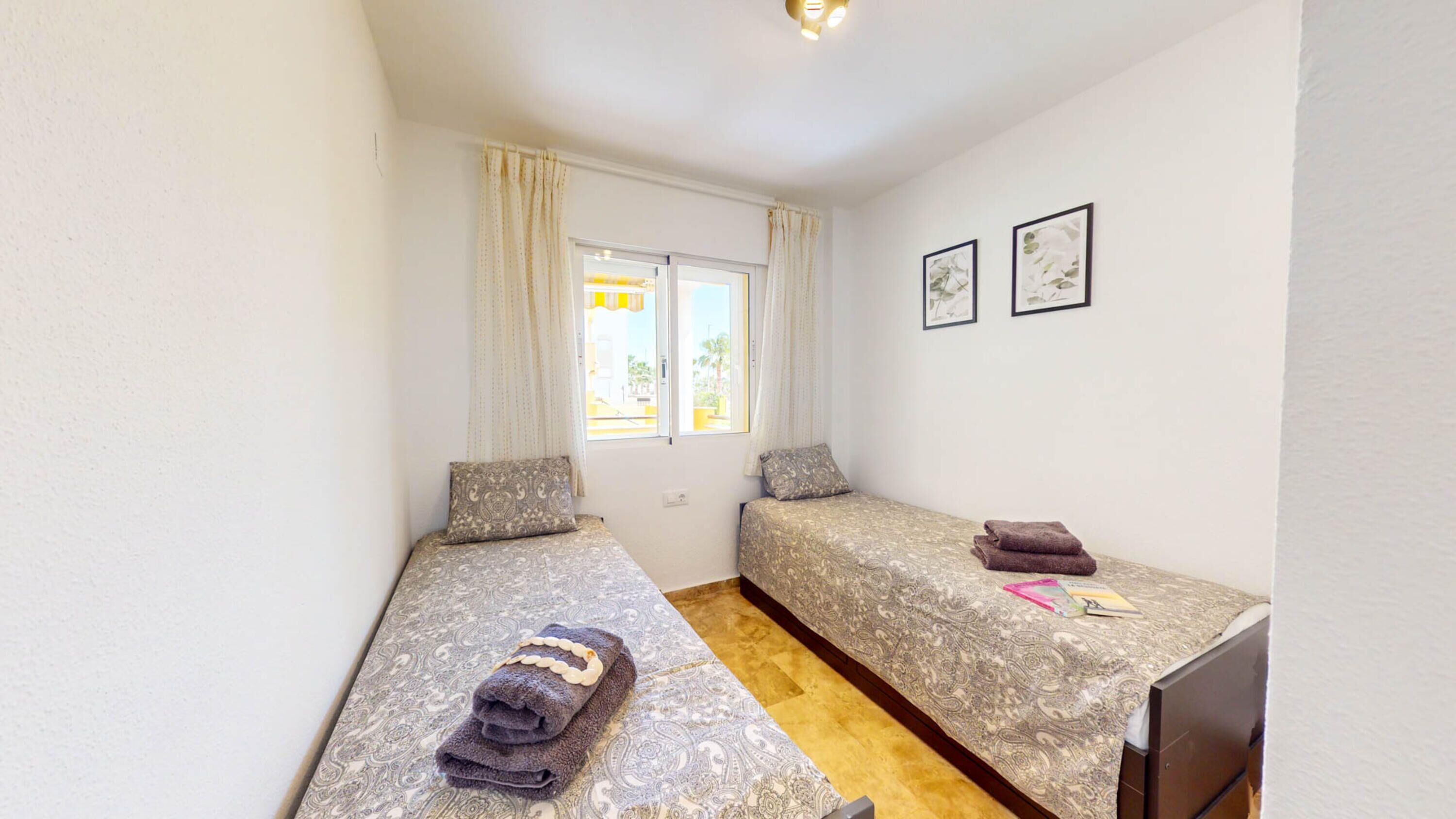 A small, well-lit room with two single beds, a window with curtains, and a desk with a chair.