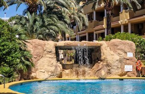 A pool with a waterfall feature, surrounded by palm trees and a building with balconies. - Apartment in Cascada de Las Marinas, 300m from the beach (Dénia)