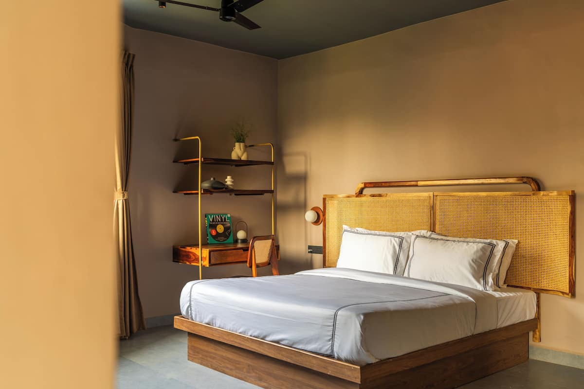 A bedroom with a wooden bed frame, a headboard with a woven design, a bedside table with a lamp, and a shelf with decorative items.