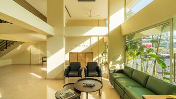 A spacious lobby with a green sofa, two black chairs, a round table with a magazine, and a large window with plants.