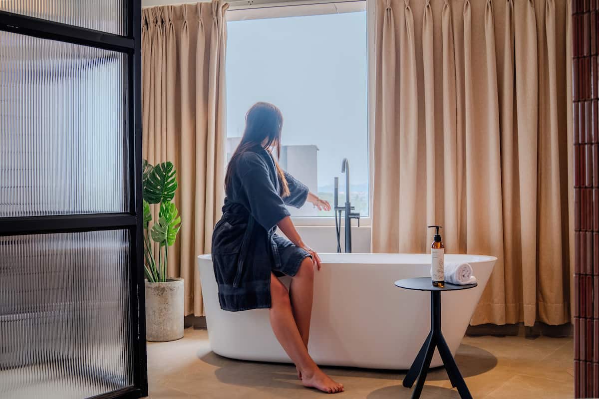 A person sitting on a bathtub in a room with a large window, a potted plant, and a small table with a bottle.