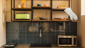 Cocina compacta con microondas, tostadora y refrigerador pequeño. Hay repisas con utensilios de cocina y un tazón de fruta sobre la encimera.