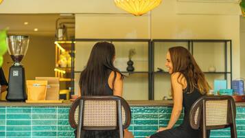 Two women seated at a counter with a green tiled backsplash, a coffee machine, and a hanging light fixture.