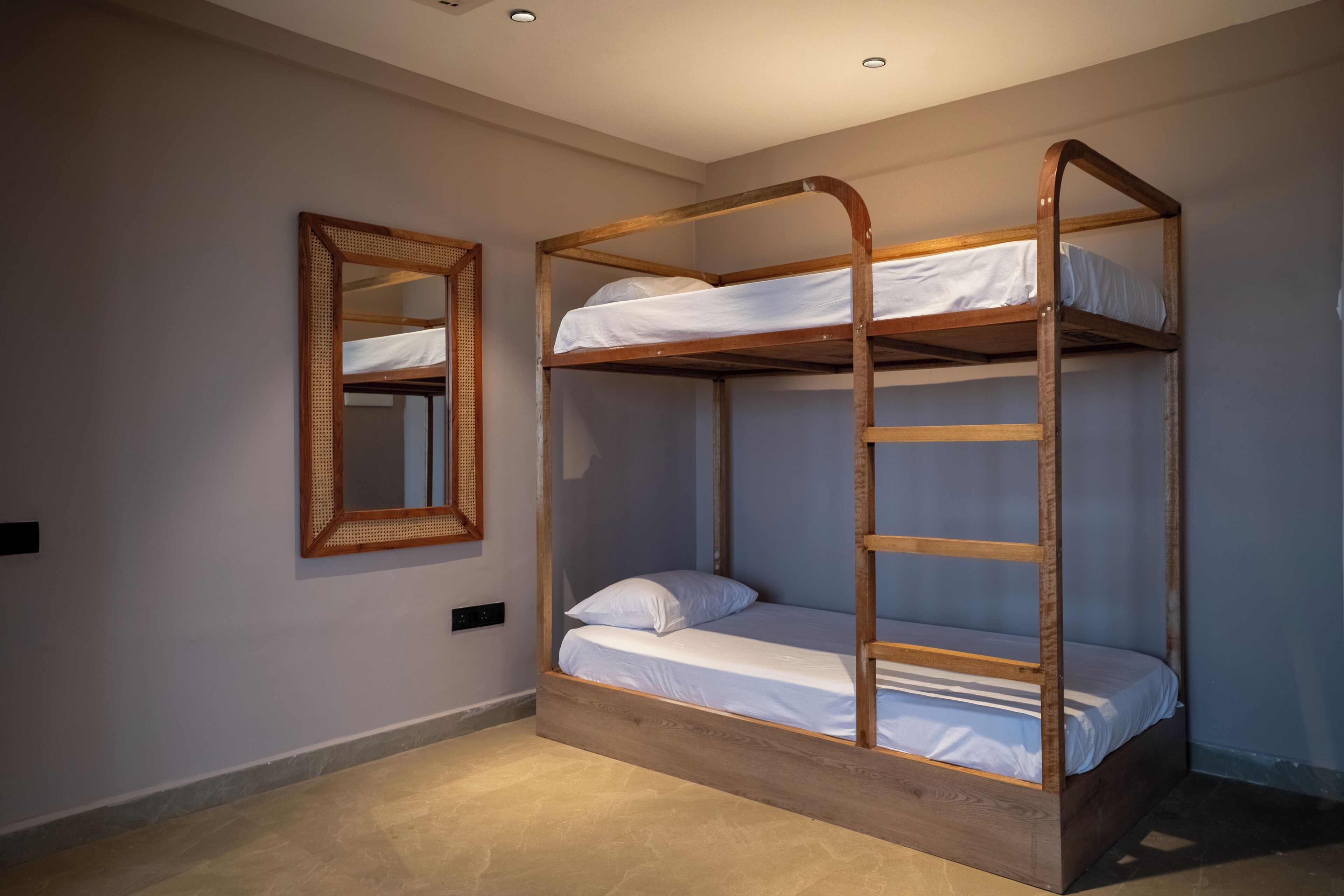 Una cama litera con estructura de madera y ropa de cama blanca, un espejo en la pared y un interruptor de luz en el suelo.