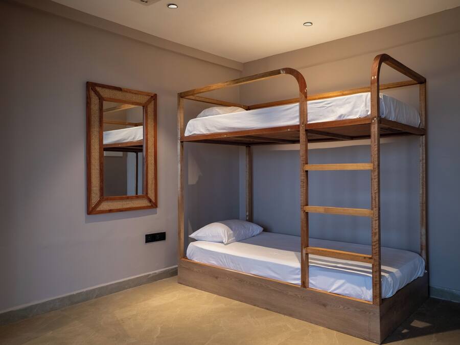 Una cama litera con estructura de madera y ropa de cama blanca, un espejo en la pared y un interruptor de luz en el suelo.