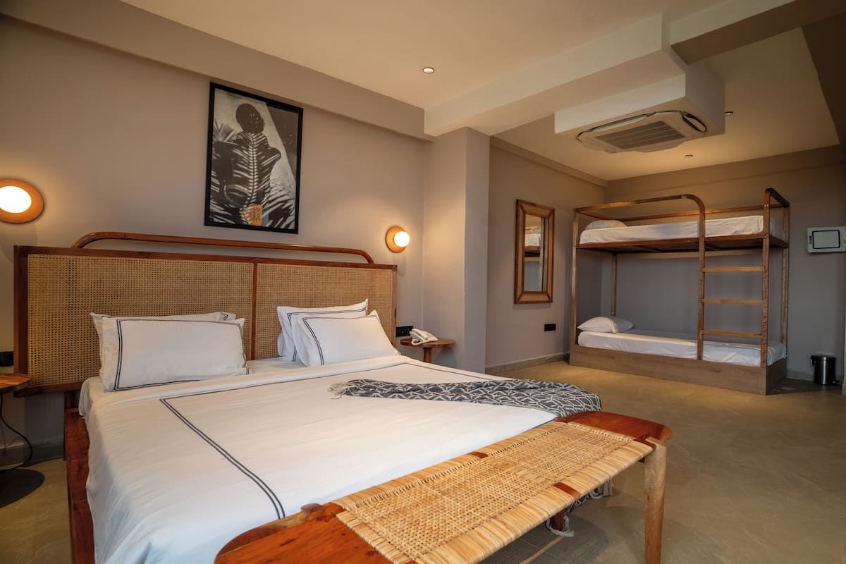 A hotel room with a large bed, a smaller bed, a bench, a bunk bed, a wall-mounted air conditioner, and a framed picture on the wall.