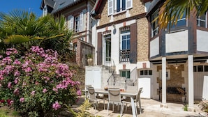 Une maison à deux étages dotée d’un patio comprenant une table et des chaises, le tout entouré d’une végétation luxuriante et de plantes en fleurs.