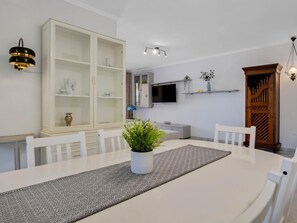 A dining area with a white table, chairs, a patterned runner, a potted plant, a cabinet with glass doors, a TV, and a wooden door. - Montemar Puerto - Beachfront, Terrace, Swimming Pool. Sea Views (Torremolinos)